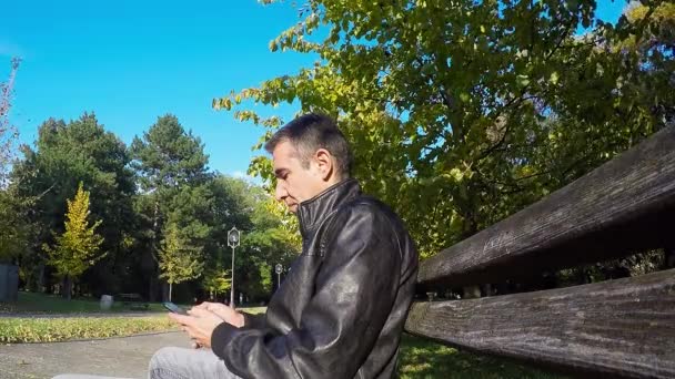 Mensajes de texto de hombre en el banco en Autumn Park — Vídeos de Stock