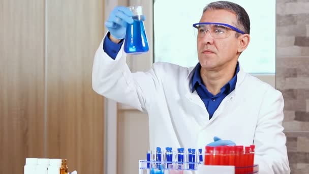 中年科学家检查一烧杯蓝色液体 科学家穿着防护装备 在实验室工作 玻璃实验室用红色和蓝色液体在白色桌上的化学试验管 — 图库视频影像
