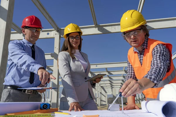 Gemengd Construction Business Team in Yellow en Red Hardhats bespreken blauwdruk op bouwplaats in aanbouw — Stockfoto