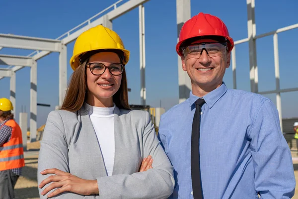 建設現場でのハードハットの男と女の笑顔 作業ヘルメットの男と女立ち サイト上のカメラを見て ビジネス ビルディング チームワークの概念 — ストック写真
