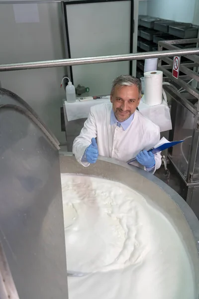 笑顔の酪農工場食品エンジニアは ステンレス鋼ミルク殺菌タンクの横に親指を上げます 白の制服と青の手袋の食品エンジニアカメラを見て 乳製品加工工場での乳殺菌 食品安全 — ストック写真