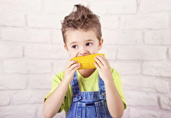 Leuke jongen eten gekookt suikermaïs op witte bakstenen muur achtergrond Rechtenvrije Stockfoto's