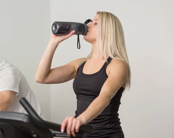女人喝水固定自行车上在健身房里 免版税图库图片