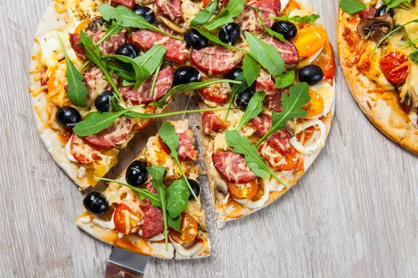 Cook takes a shovel one piece of pizza with cheese olives and salami, mushrooms and tomatoes, arugula. On a wooden stand — Stock Photo, Image