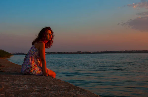 美丽的女人，看着夕阳坐在靠近水的地方 — 图库照片