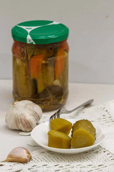 Pickled cucumber on a white plate with a fork. Canned vegetables and garlic in the background. — Stock Photo, Image