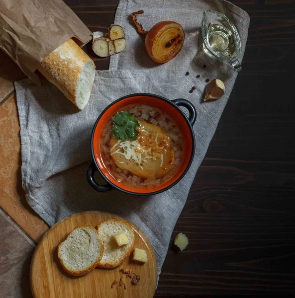 Olla con sopa de cebolla y croutons de queso en una servilleta de lino con baguette, queso sobre un fondo oscuro, vista superior — Foto de Stock
