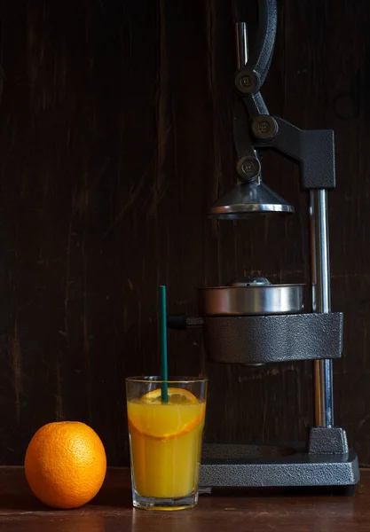 Une orange, un verre de jus fraîchement pressé avec une paille et une presse à agrumes en fonte sur fond de bois foncé — Photo