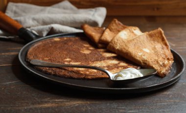 cast-iron pan with baked thin pancakes and a spoon with sour cream on a dark wooden surface, close-up