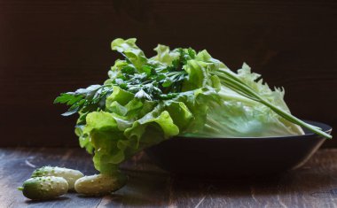 Sağlıklı bir salata için taze malzemeler, marul, maydanoz ve turşular siyah bir tabakta, ahşap arka planda.