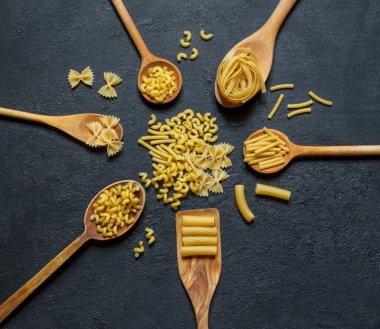 Tahta kaşıklarda farklı tipte kuru makarna siyah taş arka planda, üst açıda imza yeri var.