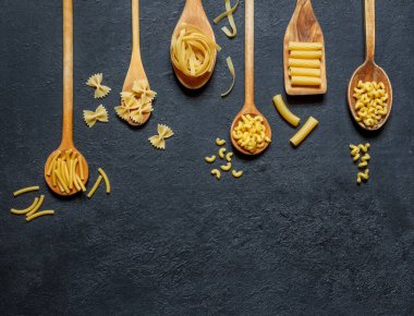 Tahta kaşıklarda farklı tipte kuru makarna, siyah taş arka planda, geniş manzaralı.