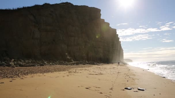 Jurské pobřeží Dorsetu v West Bay uk ráno slunce svítí na skalnaté útesy a vzdálené muž na pláži vlny — Stock video