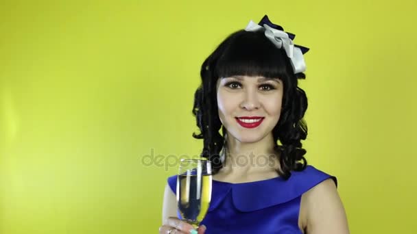 カメラの前で笑顔をシャンパンのグラスを持つ少女 黄色の背景にシャンパンを飲んで青いドレスの美しいブルネットの少女 — ストック動画