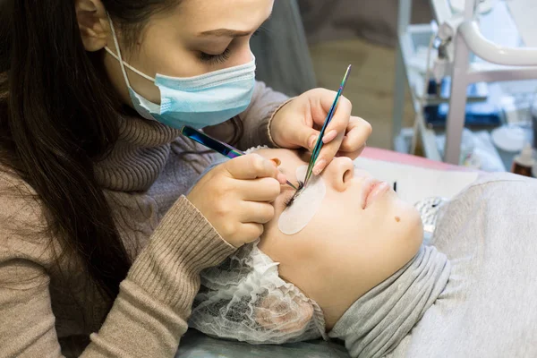 The procedure for increasing eyelashes in front of a blonde girl is done by a master in a beauty salon