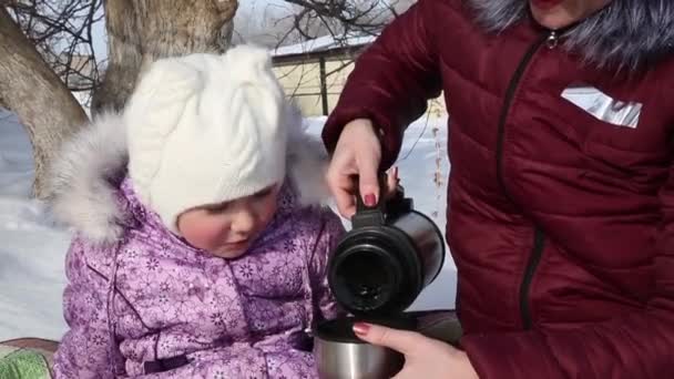妈妈和女儿在野餐时从热水瓶里喝茶 一个年轻的母亲和漂亮的小女儿坐在树下的地毯上 在冬天喝茶 — 图库视频影像