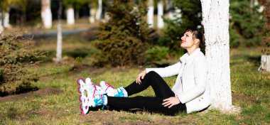 Beyaz ceketli genç esmer kadın yere oturur ve paten giyer. Paten bağlayan kız.