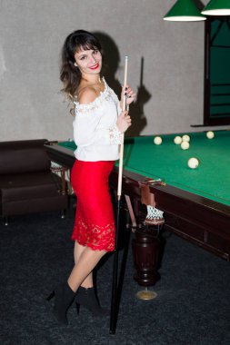 Esmer kız bilardo masasının yanında. Beyaz bluzlu siyah saçlı genç bir kadın bilardo masasının yanında elinde bir ıstaka tutuyor.
