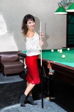 Esmer kız bilardo masasının yanında beyaz bluzlu siyah saçlı genç bir kadın kakanın yanında duruyor.,