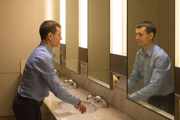 A man washes his hands in a public toilet. A 35-40 year old man is washing himself in the toilet looking at himself in the mirror