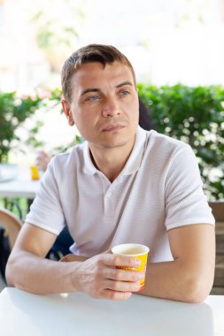 35-40 yaşındaki bir adam, bir sokak kafesinde elinde bir bardak kahveyle oturan yalnız ve üzgün bir adamdır. Yalnız bir adam bir yazlık kafede oturuyor..