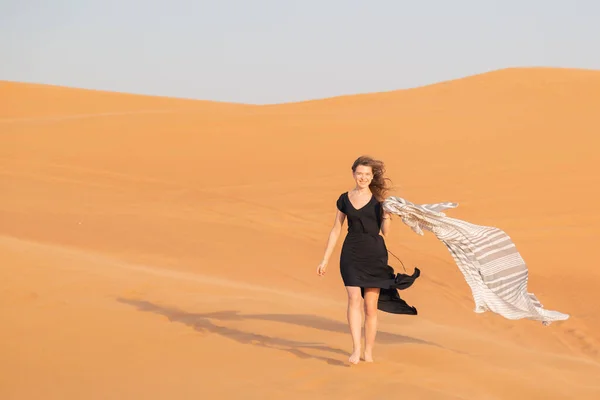 A girl walks on the sand in a deserted desert. A woman in a black dress with a cloth in her hands in strong wind alone in the desert.