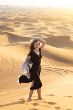 Kız model çölde fotoğrafçıya poz veriyor. 20-25 yaşlarında, siyah elbiseli, sıcak çölde dans eden beyaz bir kadın..