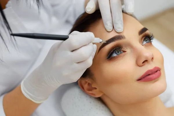 Kosmetikerin macht Augenbrauen-Make-up-Tätowierung auf Frau mit Maschine — Stockfoto