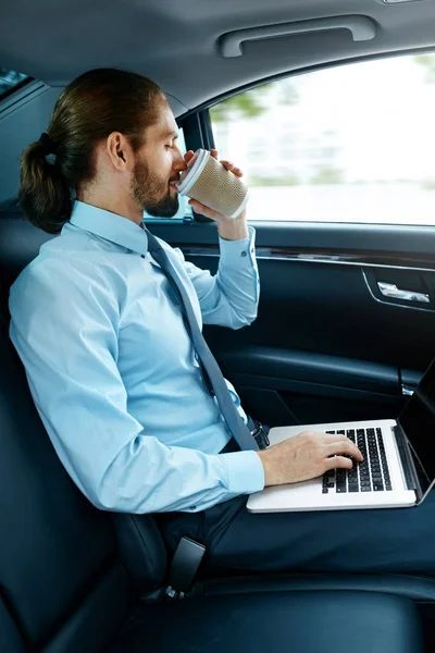Человек работает над ноутбуком и пьет кофе в машине . — стоковое фото