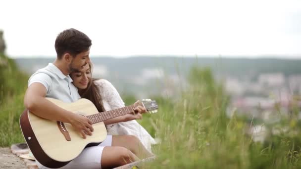 一緒に時間を過ごすロマンチックな日に自然の中の愛の美しいカップル ハンサムな若者に音楽を楽しむ美しい笑みを浮かべて女性ながらギターを弾く 公園でのピクニックの人々 — ストック動画