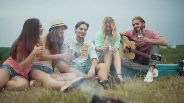 Amigos felices bebiendo cerveza, divirtiéndose y sentados cerca de la hoguera — Vídeos de Stock