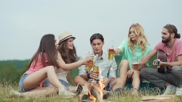 Amigos felices bebiendo cerveza, divirtiéndose y sentados cerca de la hoguera — Vídeos de Stock
