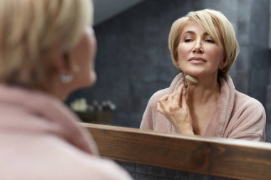 Cilt rutini. Olgun kadın, aynanın yansımasının önünde, yüz derisi üzerinde Jade Roller kullanır. Yaşlanma Karşıtı Güzellik Tedavisi Konsepti.