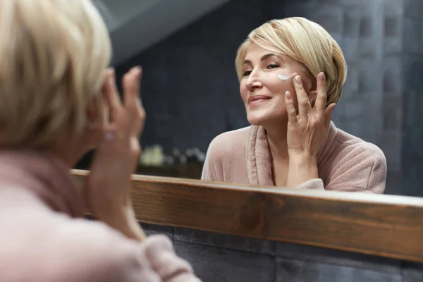 スキンケアルーチン 熟女は鏡面反射の前で顔の皮膚に化粧品クリームを使用しています アンチエイジング美容トリートメントコンセプト — ストック写真