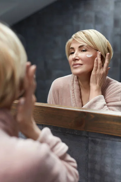 スキンケアルーチン 熟女は鏡面反射の前で顔の皮膚に化粧品クリームを使用しています アンチエイジング美容トリートメントコンセプト — ストック写真