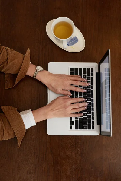 女性与笔记本电脑 妇女的手穿着棕色夹克在办公桌前的笔记本上打字 一杯茶 计算机概念的使用 — 图库照片