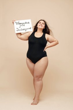 Artı Beden Modeli. Siyah elbiseli şişman kadın boy portresi. İyi göründüğünü düşündüğün zaman boş bir kağıtta yazan kadın kâğıdı elinde tutuyor. Gülümseyen Vücut Pozitif Esmer.