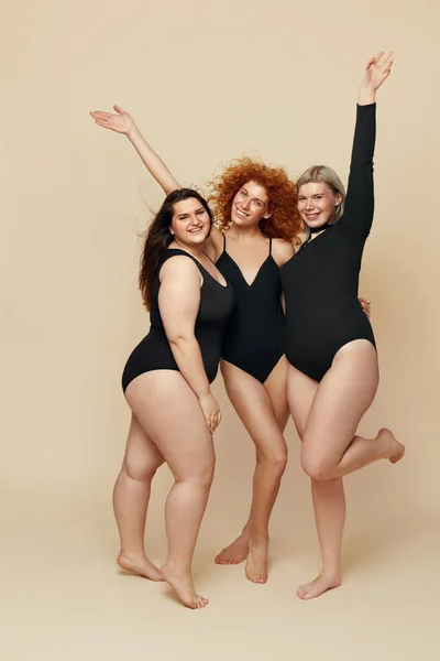 Different Body Types. Group Of Diversity Models Full-length Portrait. Blonde, Brunette And Redhead In Black Bodysuits Posing On Beige Background. Female Friendship For Happy Life.