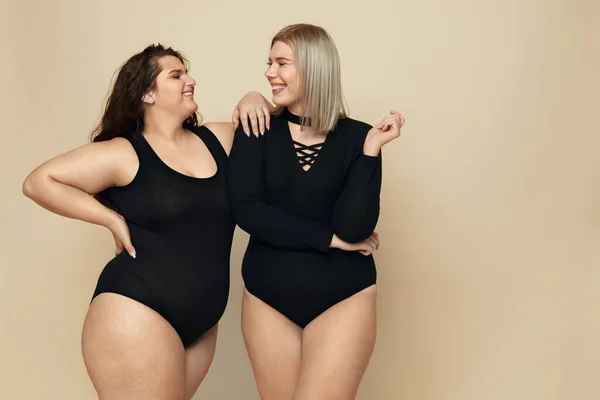 Plus Size Models. Full-figured Women Portrait. Smiling Brunette And Blonde In Black Bodysuits Posing On Beige Background. Body Positive As Lifestyle.