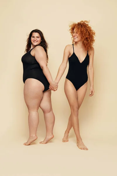 Different Size. Diversity Women Full-Length Portrait. Smiling Brunette And Redhead In Black Bodysuits Posing On Beige Background. Female Friendship.