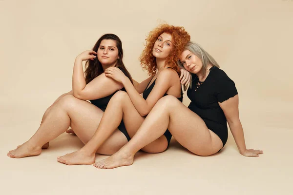 Different Body Types. Diversity Models Portrait. Group Of Women Posing On Beige Background. Blonde, Brunette And Redhead In Black Bodysuits Sitting On Floor.