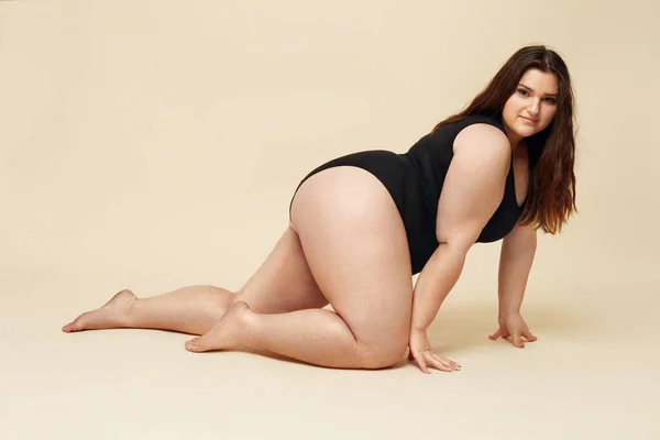 Plus Size Model. Fat Woman In Black Bodysuit Portrait. Crawling Brunette Posing On Beige Background And Looking At Camera. Body Positive Concept.