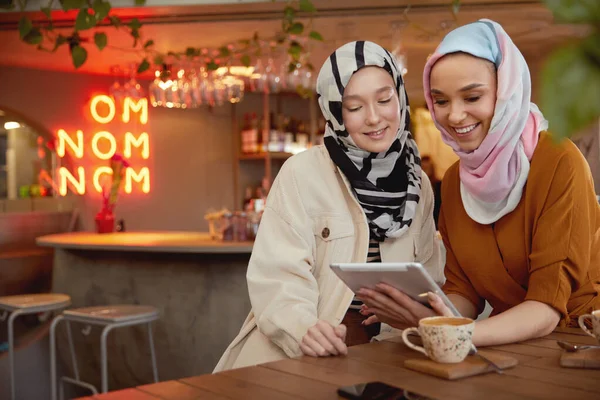 Women. Young Girls In Hijab. Friends Meeting In Cafe. Smiling Muslim Female Drinking Coffee And Looking On Tablet.