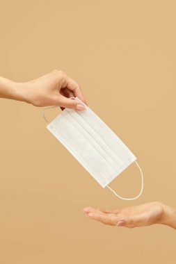 Korunma. Enfeksiyon önleme maskeli eller. Beyaz Maskeli Kadın, Bej arka plandaki birine Beyaz Maske Veriyor. Sağlık hizmetleri için Tıbbi Malzeme Kullanımı.