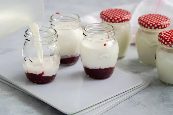 Filling glass jars with a milk mixture to produce homemade yoghurt. Raspberry jam at bottom of jars to add a delicious sweet flavor. Organic dairy product, healthy eating and sustainable concept