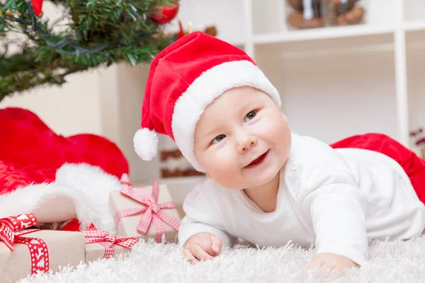 Bébé mignon dans un chapeau de Père Noël à côté de l'arbre de Noël avec des cadeaux — Photo