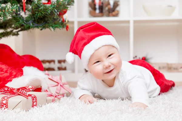 Bébé mignon dans un chapeau de Père Noël à côté de l'arbre de Noël avec des cadeaux — Photo
