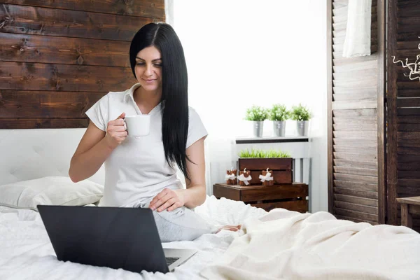 Relaxează-te pe pat. O femeie frumoasă se bucură de o ceașcă de cafea și își folosește laptopul — Fotografie, imagine de stoc