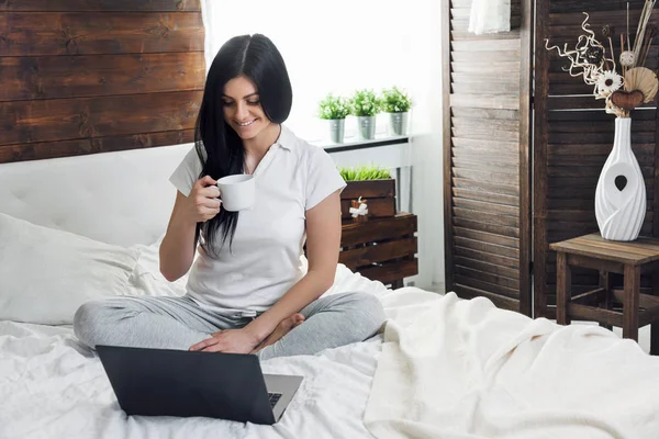 Relaxar na cama. Mulher bonita desfrutando de uma xícara de café e usando seu laptop — Fotografia de Stock