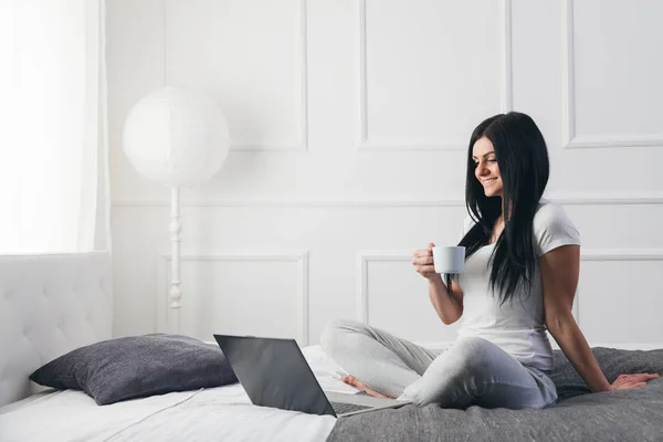 Ontspannen op het bed. Mooie vrouw genieten van een kopje koffie en het gebruik van haar laptop — Stockfoto